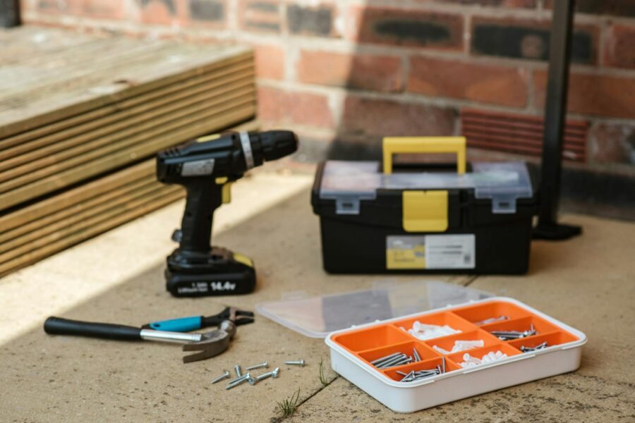 Electric screwdriver on floor near containers with instruments placed near spanner with hammer against brick wall and wooden barrier