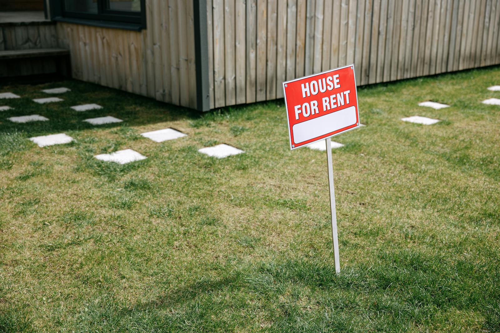 A red 'House for Rent' sign stands on a grassy lawn beside a wooden house exterior.