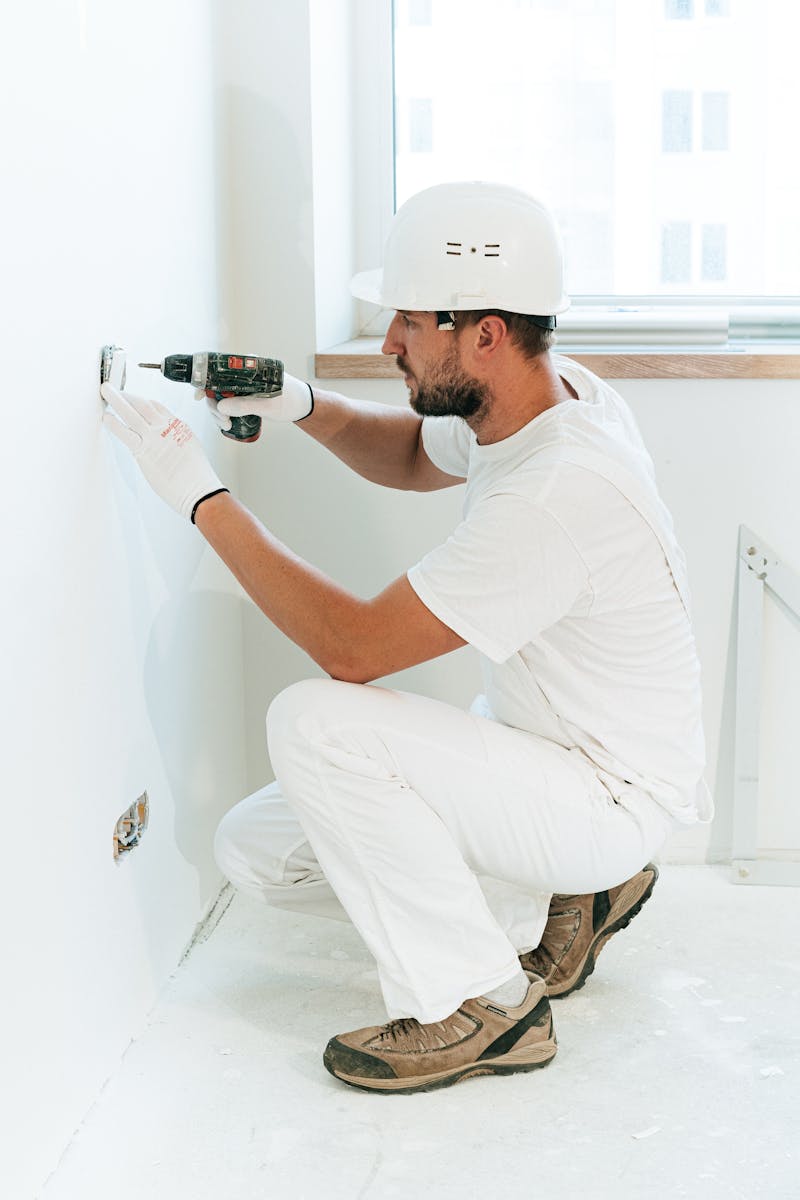 A construction worker wearing a helmet uses a drill to install a wall outlet inside an apartment.