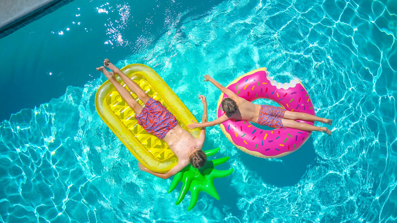 Two boys sunbathing on colorful pool floats, enjoying a sunny day in a clear blue swimming pool.