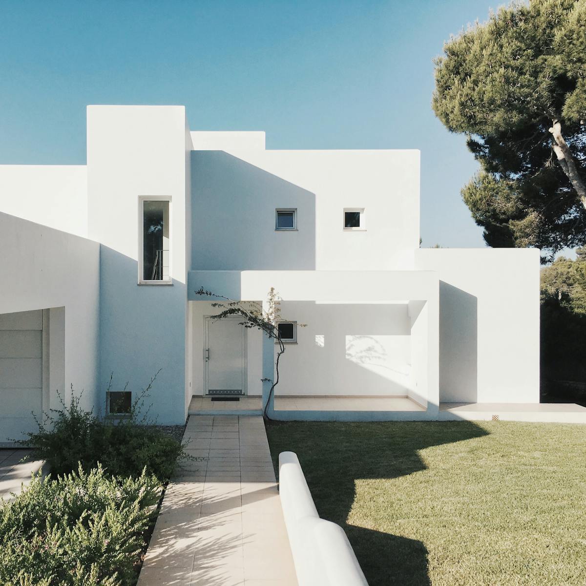 A minimalist modern white house exterior with clean lines and geometric shapes, set against a clear blue sky.