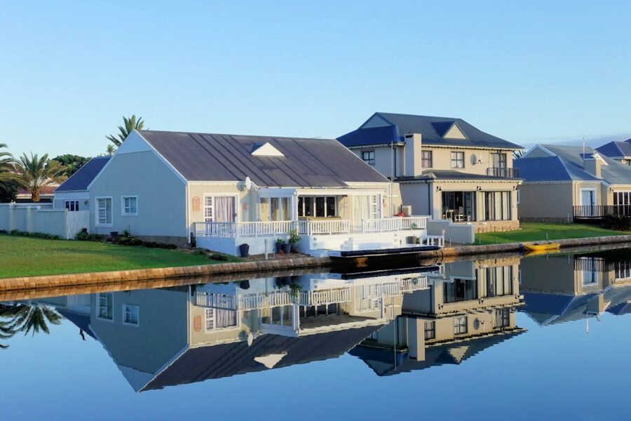 Luxury modern homes reflecting on a peaceful, clear lake during daylight, showcasing architectural beauty.